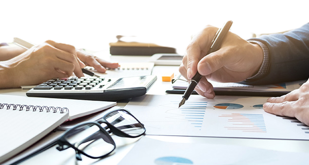 image focused on the hands of two people working accross from eachother. One is entering numbers in a calculator the other is writing. Both persons are sitting at a table across from eachother.